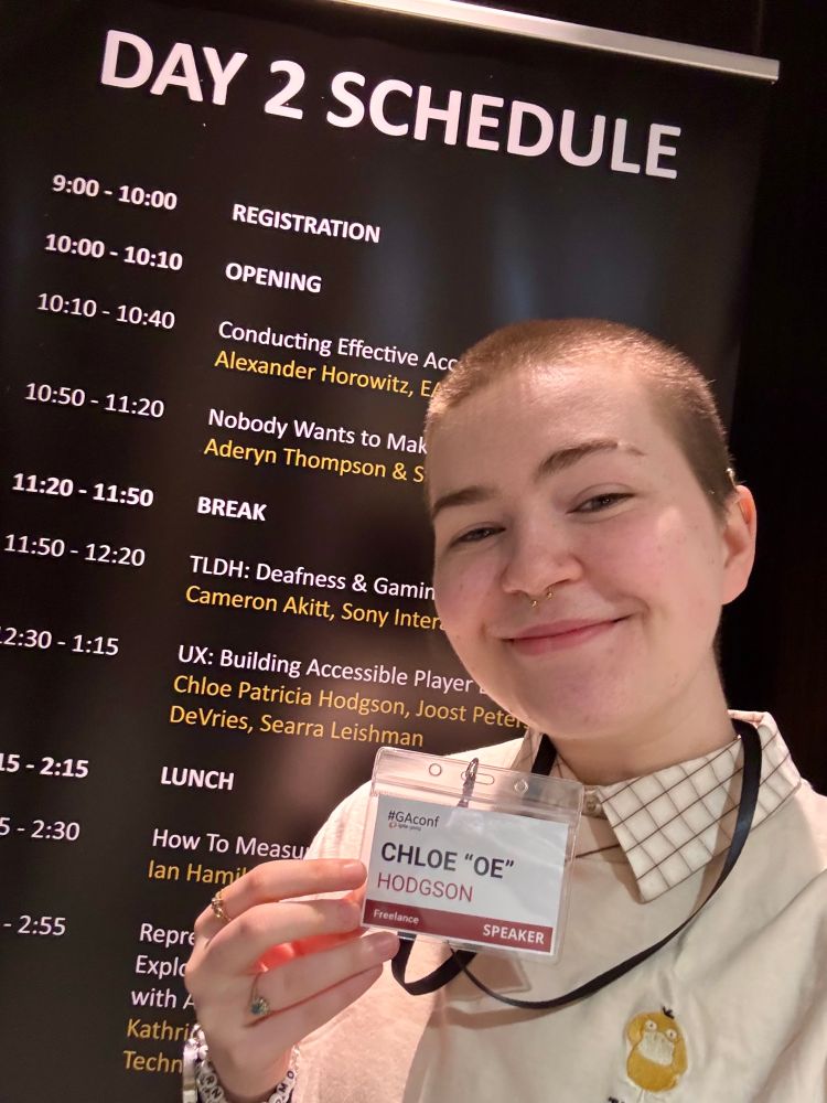 a smiling selfie with the talk schedule, holding my pass with my name on it. I am young, white and androgynous, with a shaved head, and wearing a shirt with a psyduck on it