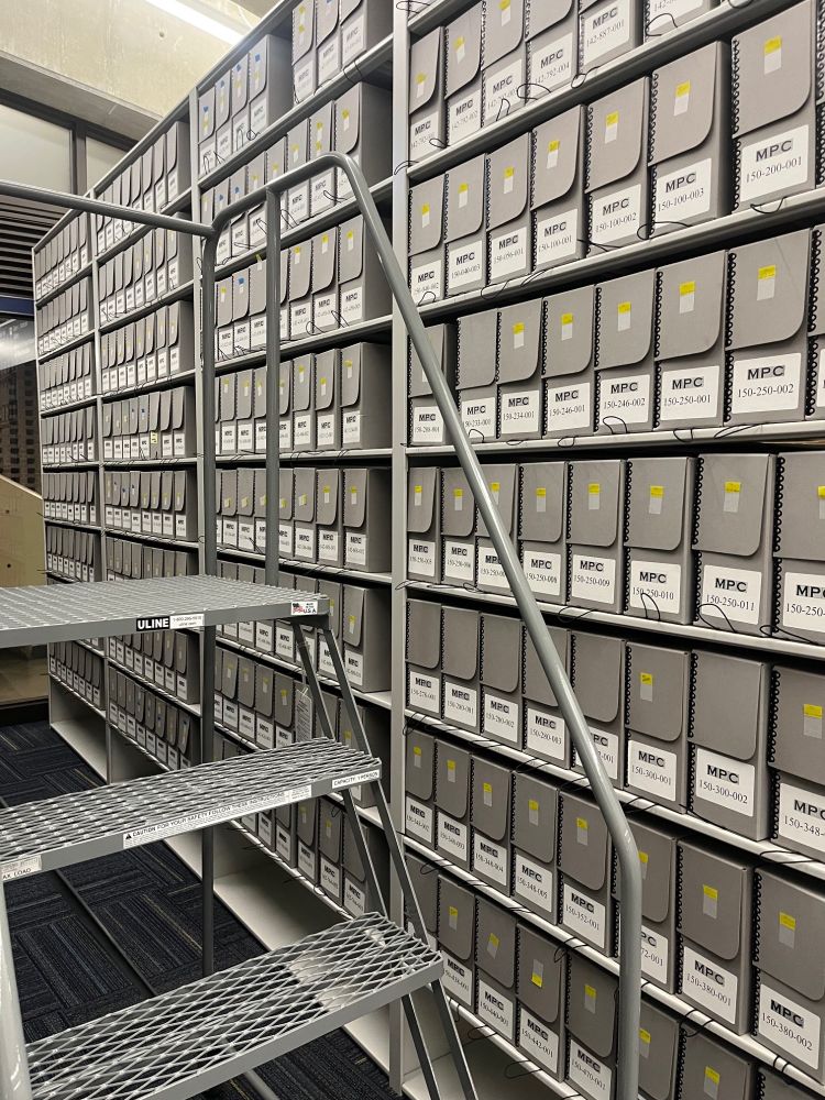 A movable metal staircase is in front of a tall set of metal shelved filled with small, vertical document boxes all neatly labeled and securely housed in IPUMS HQ. 