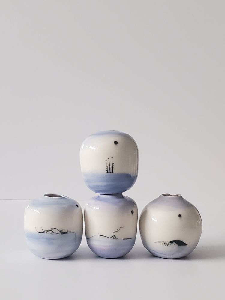 Four stacked, small, and porcelain vases. Three on the bottom and one on top. They are on a white background. All have light blue or purple bottoms and tops. The middles are decorated with black underglaze.

Bottom row

Vase 1: the scene is on a  small hill with wisps of grass on either side

Vase 2: the scene is of a small triangular house on a hill with smoke coming from a tiny chimney.

Vase 3:  Top vase 2: a small setting sun painted black with housing structures next to it.

The design of the top vase includes three segmented reeds next to each other with a dot at the top of each reed. 

 

