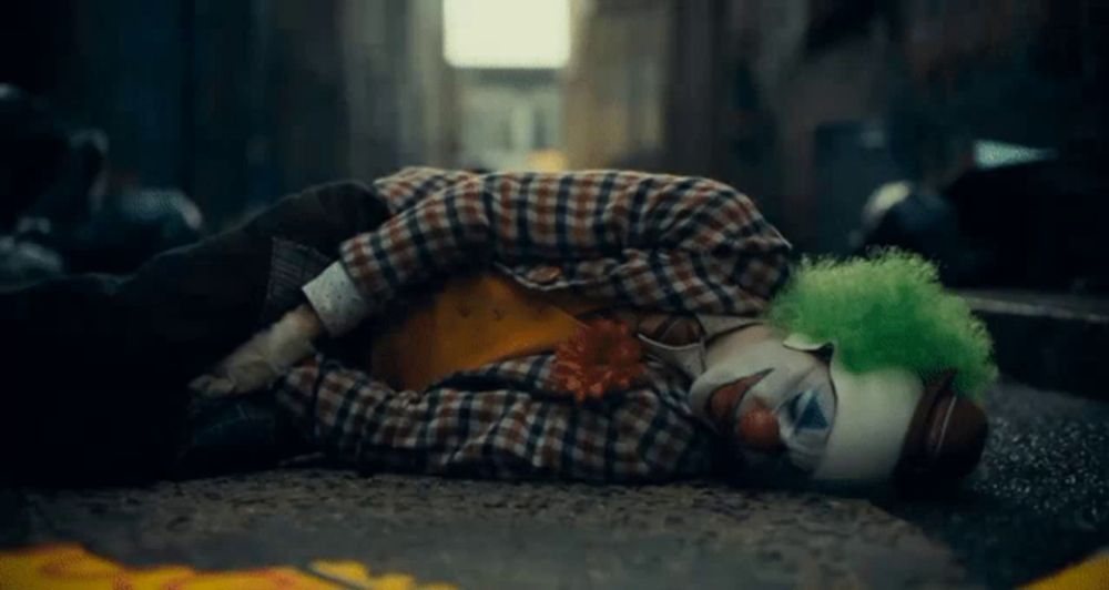 the joker is laying on the ground in a plaid shirt and green clown wig .