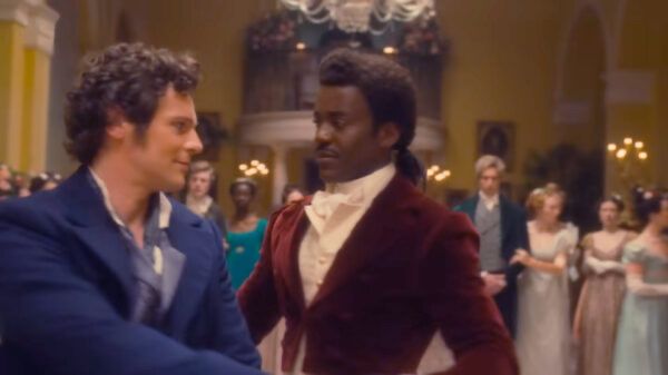 A still photo from the current season of Doctor Who: episode title “Rogue” which premiered on June 7 of the Doctor and another character played by Jonathan Groff dancing together in a ballroom. They are in regency era costumes and behind them, an audience also in regency era costumes looks on.