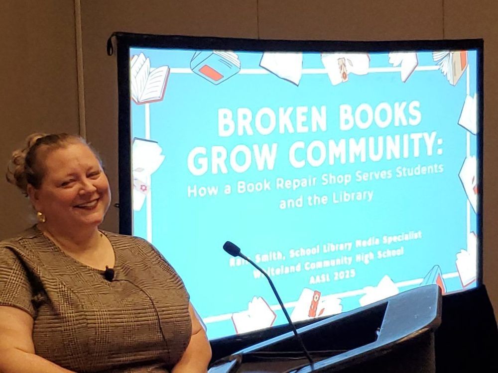 Person sitting and smiling to the side in front of a presentation screen titled 'BROKEN BOOKS GROW COMMUNITY: How a Book Repair Shop Serves Students and the Library,' 