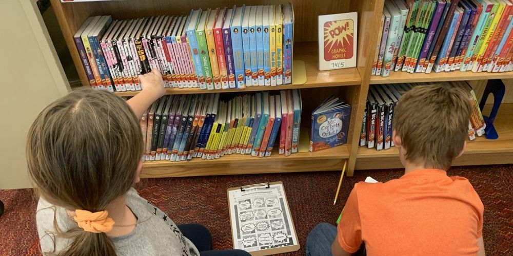 Two children in a library, one selecting a book from a shelf filled with colorful children's books including titles like "Bone" by Jeff Smith, while the other is seated on the floor looking at a comic book.