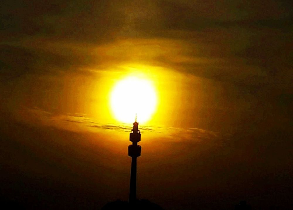 Die gelb umrandete Sonnenscheibe strahlt durch eine dunklen Wolkenvorhang, in dem Moment fotografiert, an dem sie genau über der schwarzen Silhouette eines Fernsehturms steht 
