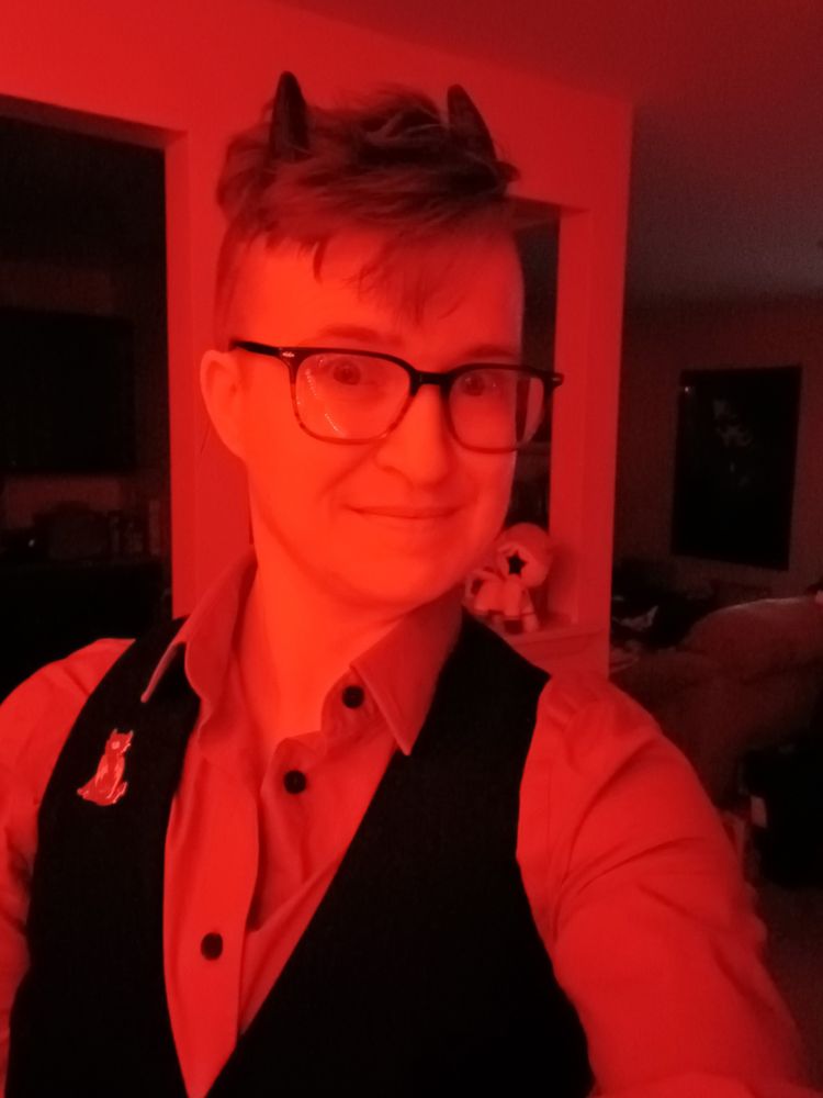 a short haired nonbinary person with small devil horns bathed in red light