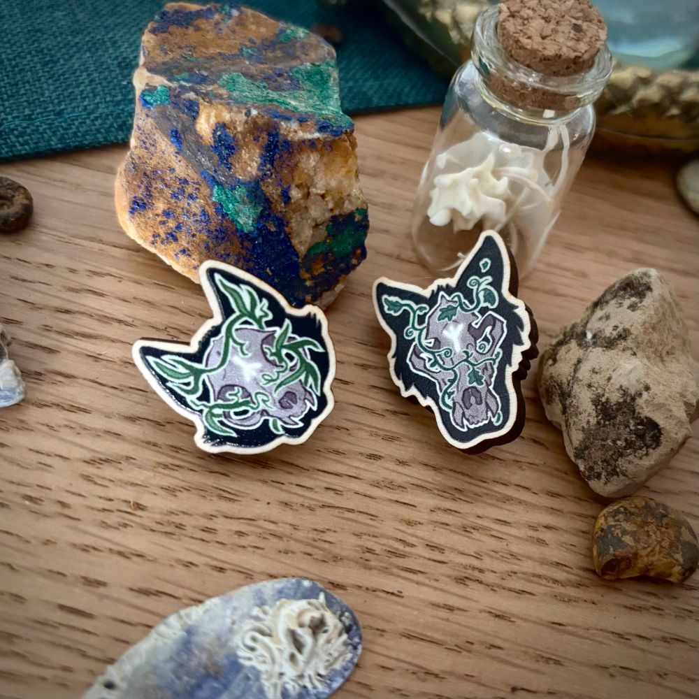 Photo of two wooden pins, a cat skull and dog skull, overgrown with vegetation, emblazoned with a rune