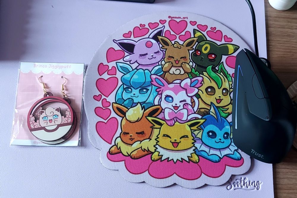 Foto do brinco da Jigglypuff dentro de uma pokébola, que só mostra a parte de baixo. No fundo, tem flores de sakuras. O brinco é de MDF. Do lado, o mousepad com todas as eeveelutions, com Flareon, Jolteon e Vaporeon na base; Glaceon, Sylveon e Leafeon no meio; e Espeon, Eevee e Umbreon em cima. Todos estão cercados de corações rosa.