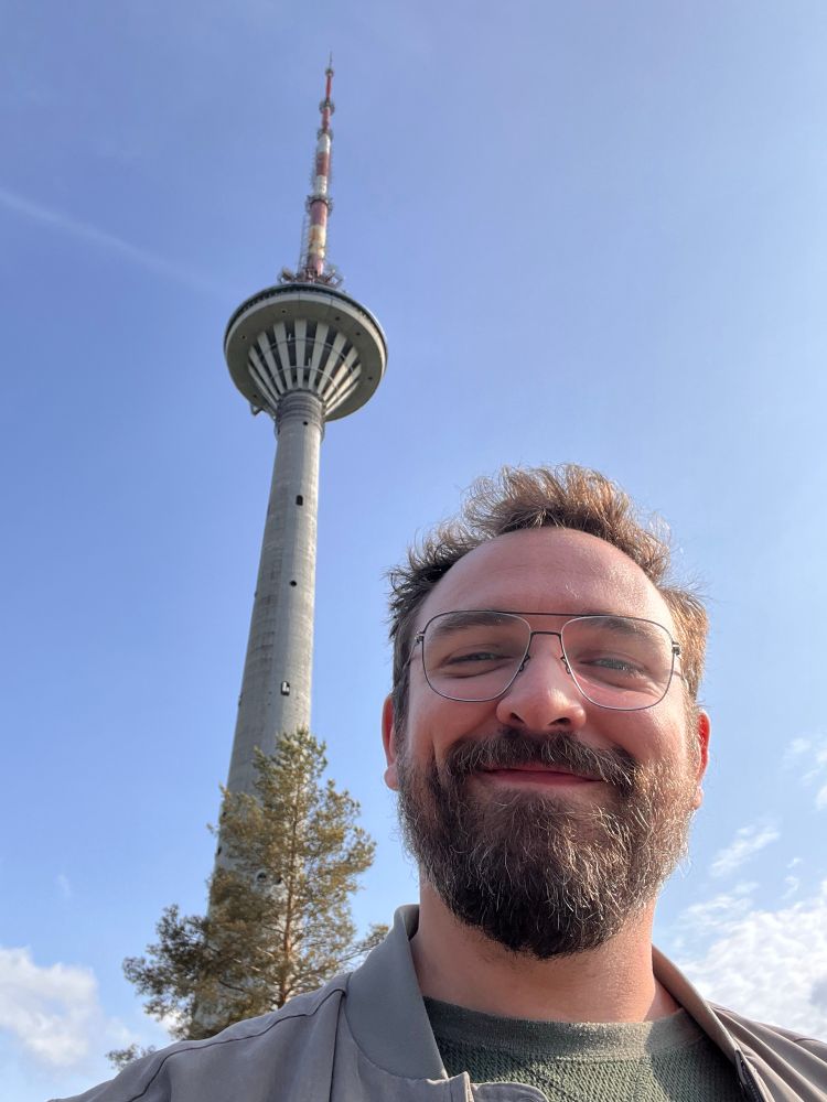A man with glasses wearing a green shirt and green jacket is smiling in front of a TV Tower