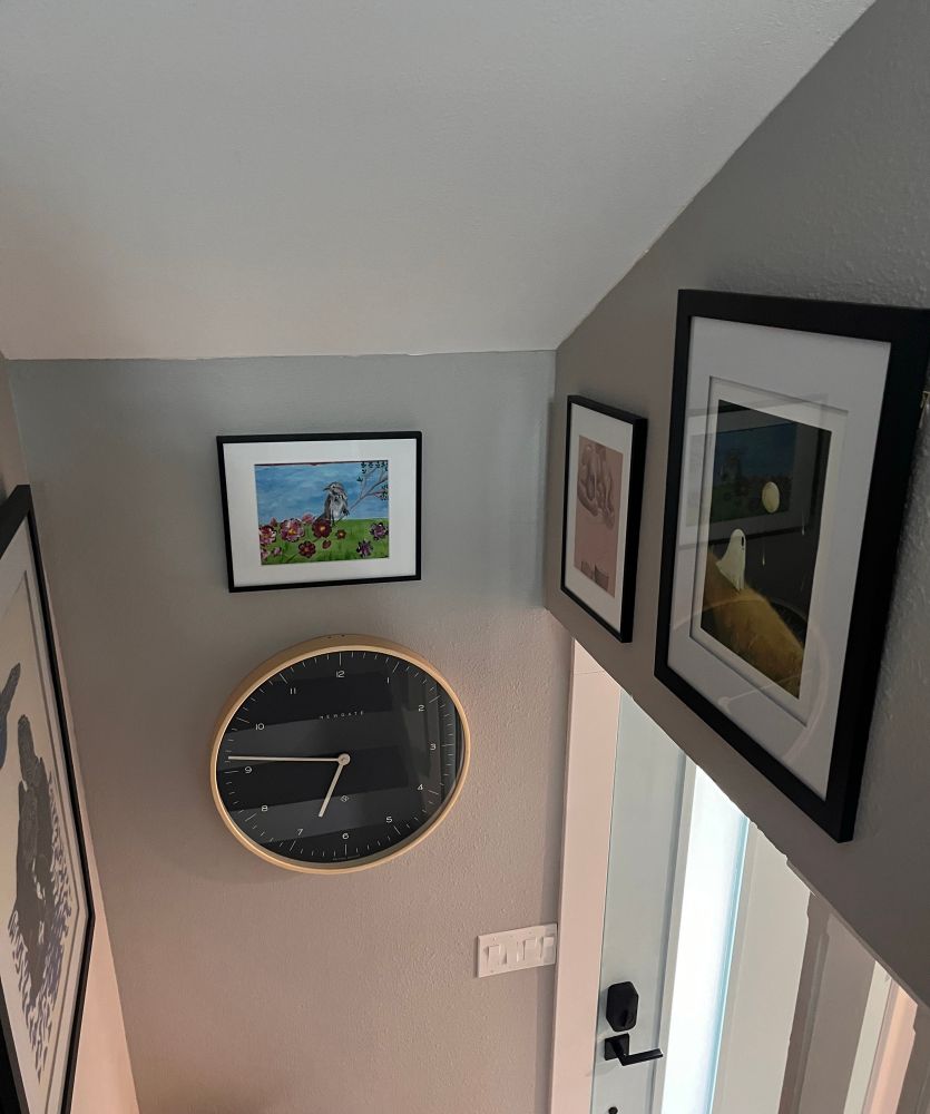 Art work in black frames of varying sizes share a wall with a large clock next to a door