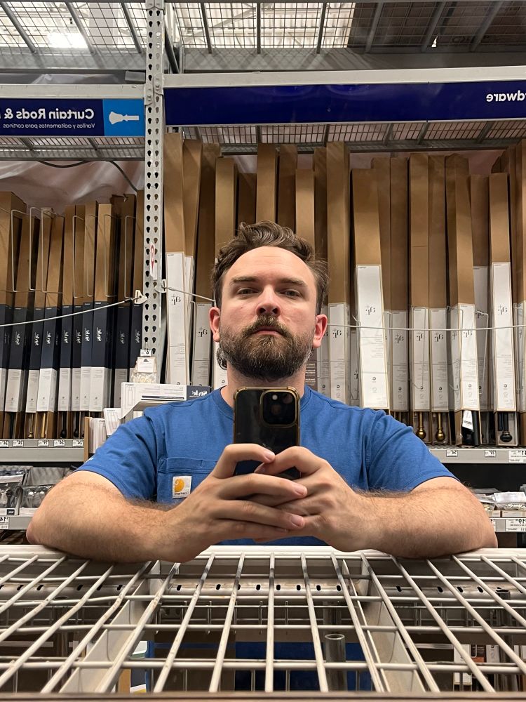 A man takes a mirror selfie in the Curtain Rod section of Lowe’s

He is wearing a blue shirt