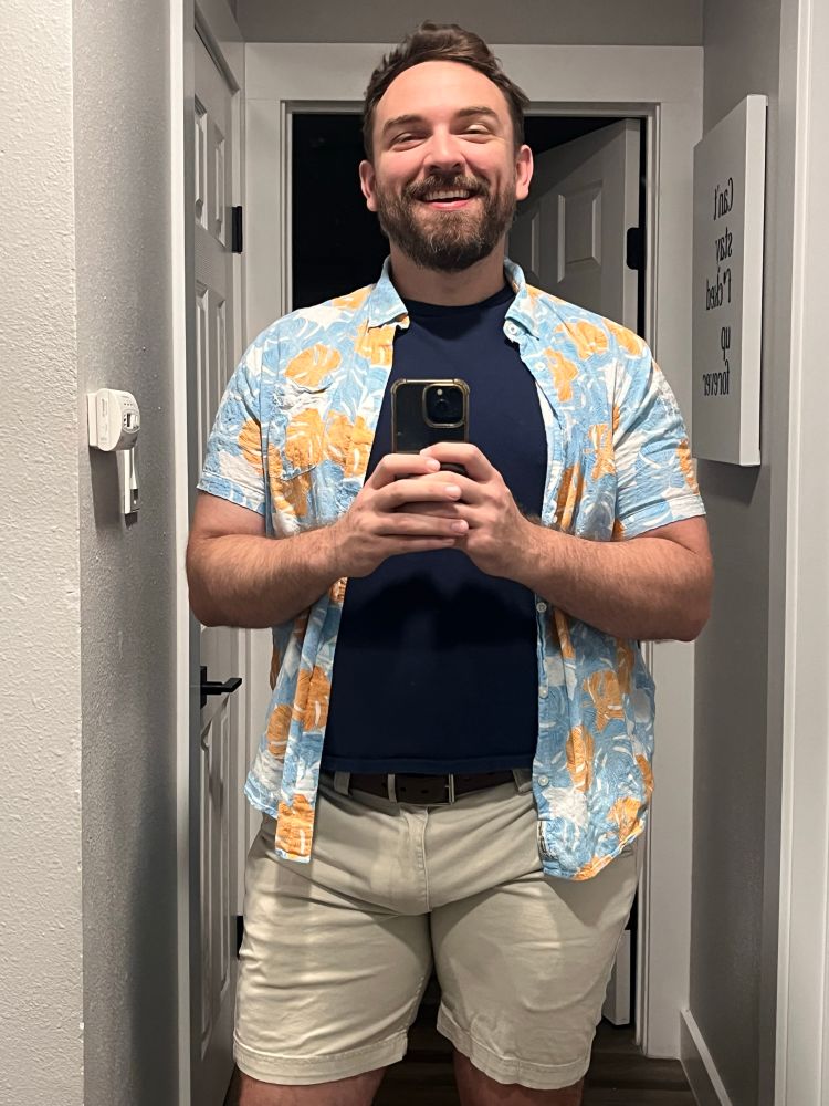A man is smiling while taking a mirror selfie

He is wearing white shorts a brown belt a dark blue shirt and a tropical overshirt