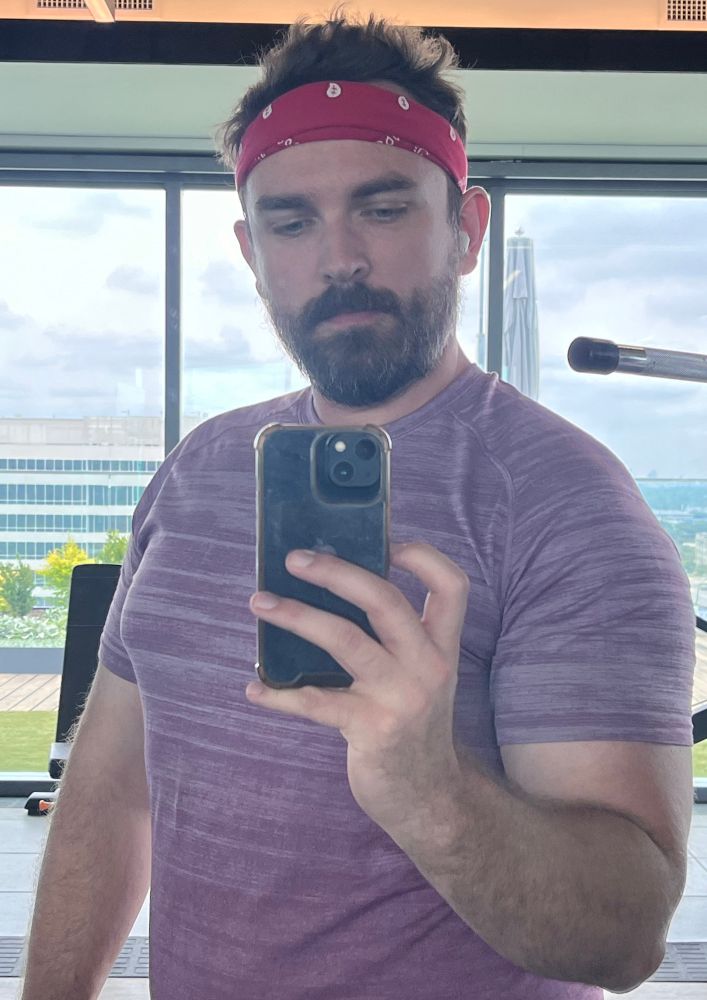 A man takes a mirror selfie while wearing a red bandana and a purple workout shirt
