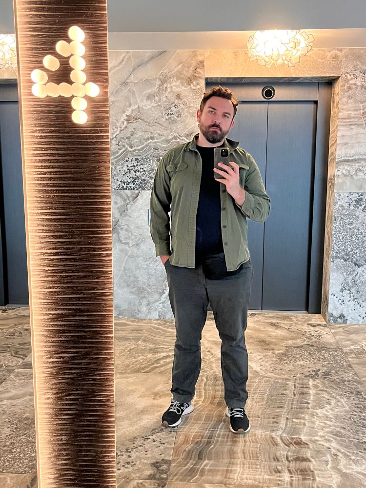 A man takes a mirror selfie in front of an elevator/lift door - he’s wearing a dark shirt and green overshirt with grey pants and untied shoes