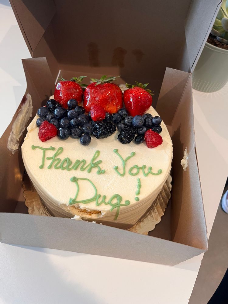 A berry covered cake in a box. The cake has the words - Thank You Dug - written on the cake