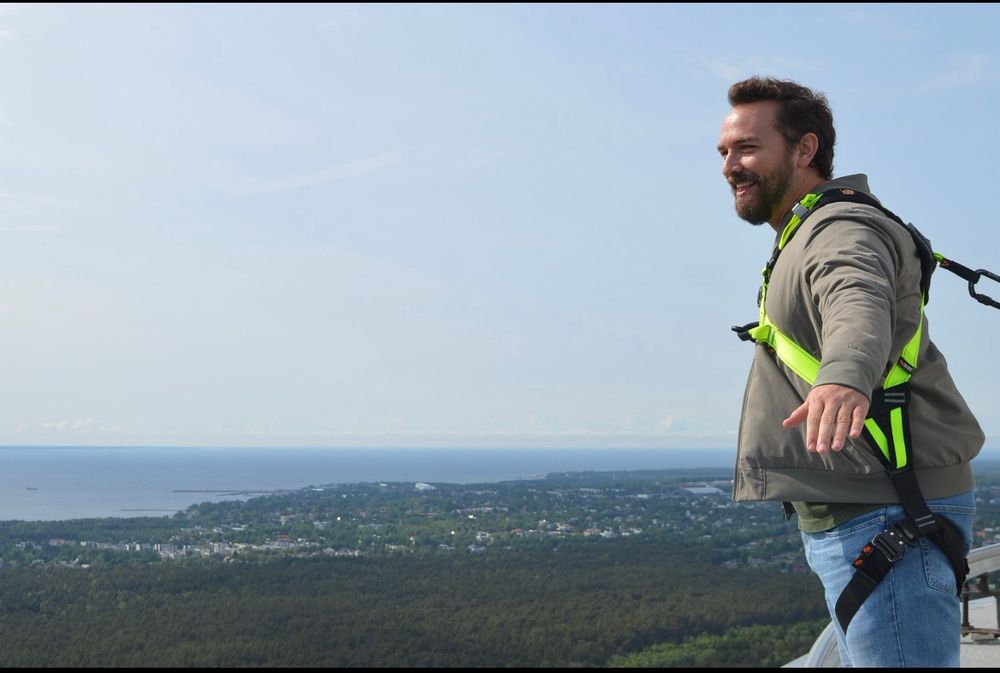 A man wearing a green jacket and jeans with a neon green body harness leans out over the edge of a sky tower

A view of forest and ocean is in the distance
