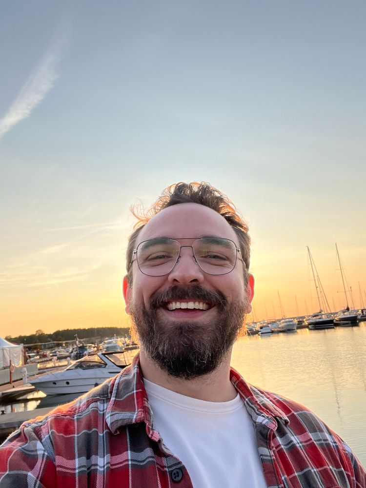 A bearded man wearing glasses, a white shirt, and a flannel smiles while taking a selfie in front of boats on water as the sun is setting