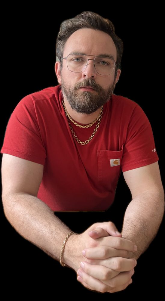 A bearded man is wearing glasses, a red shirt, a gold bracelet, and two gold chains