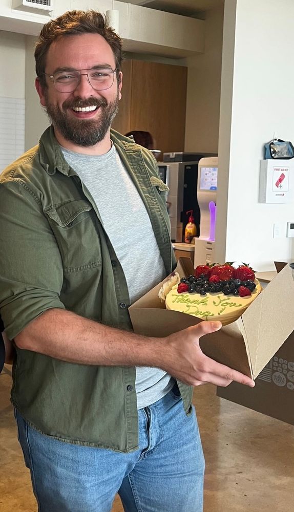 A man wearing glasses, a green overshirt, a grey tshirt, and blue jeans is standing in a kitchen while holding a cake with berries and writing on it