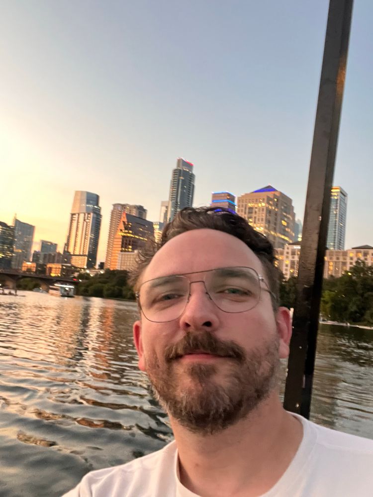 A man wearing glasses in a white shirt takes a selfie in front of the Austin skyline