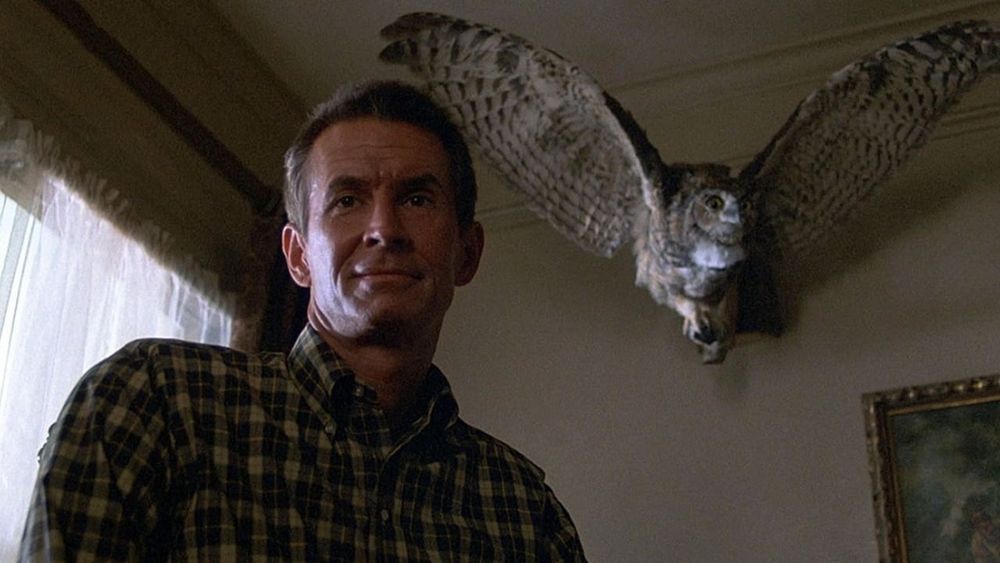 Norman Bates (Anthony Perkins) smiles in the corner of a room with a stuffed owl to his right and a window to his left in Psycho III (1986)