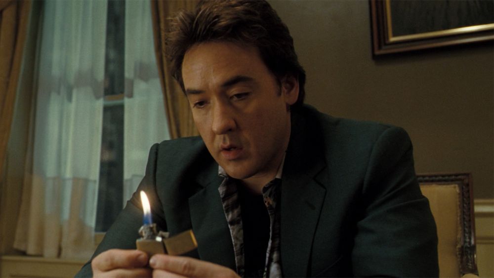 Mike (John Cusack) sits in a hotel room with a window behind him looking at an open, lit Zippo in 1408 (2007)