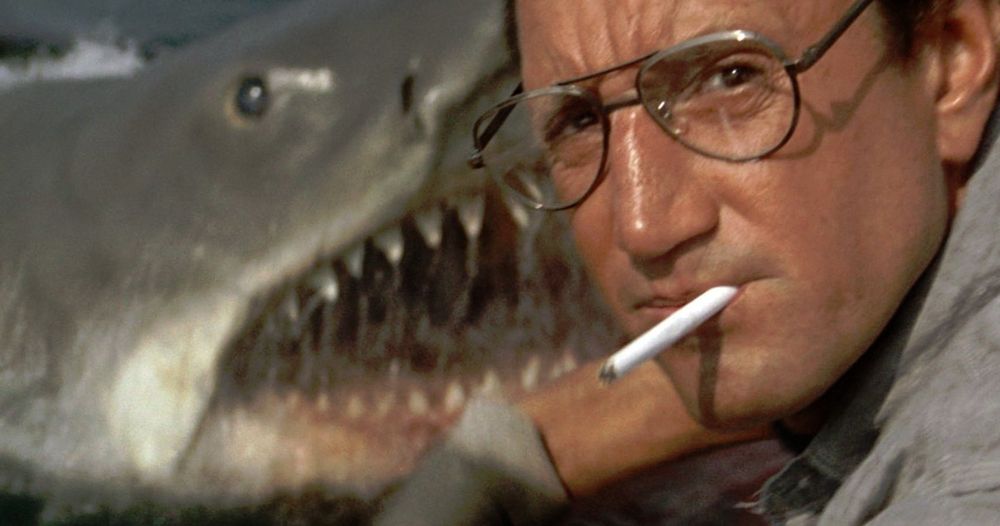 Martin Brody stands in the foreground looking at the camera with a cigarette in his mouth while the titular shark pops up in the background in Jaws (1975)