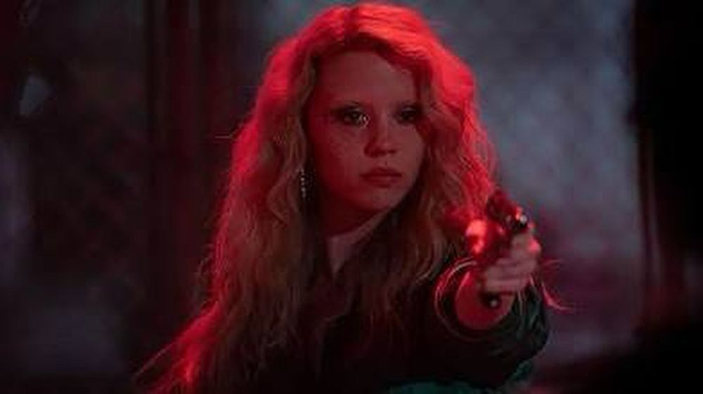 Maxine Minx (Mia Goth) holds a gun out in front of a chain link fence in Maxxxine (2024)