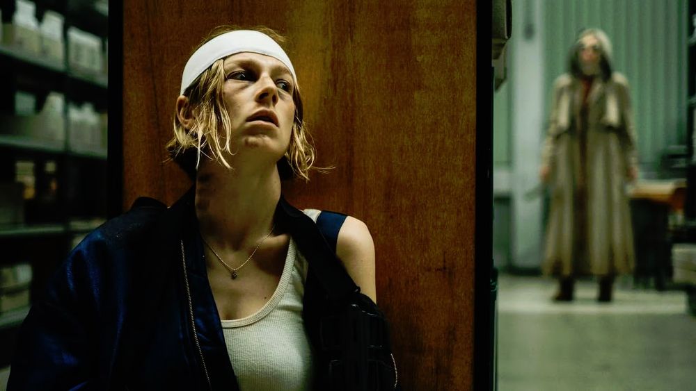Gretchen (Hunter Schafer), wearing a bandage on her head, and with her arm ina  sling, hides behind a bookcase while the Hooded Woman (Kalin Morrow) is in the background in Cuckoo (2024).