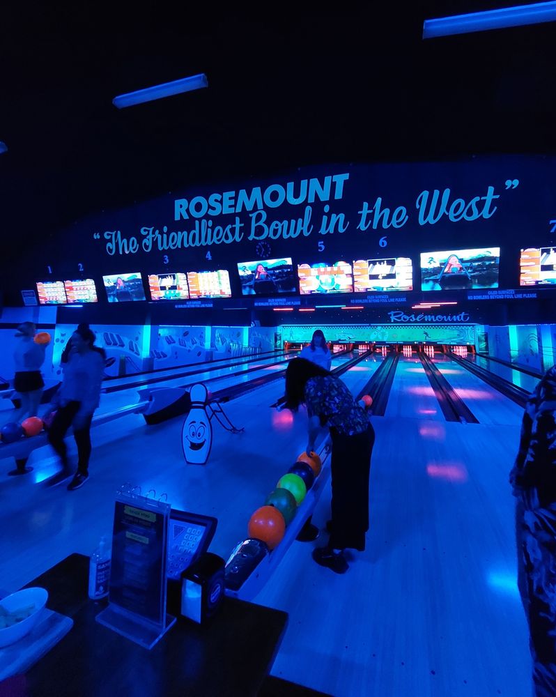 A ten pin bowling alley in the dark with blue lighting. The sign over the lanes says " ROSEMOUNT. The friendliest bowl in the west".
