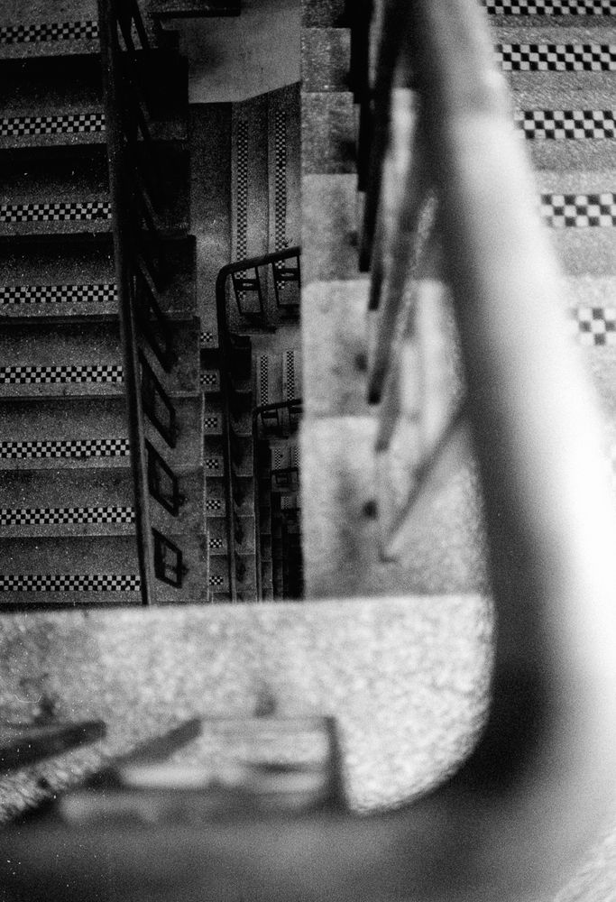Looking down the middle of a stack of stairwell banisters. The banisters are iron and the treats of the steps have tiny black and white chequered tiles. 