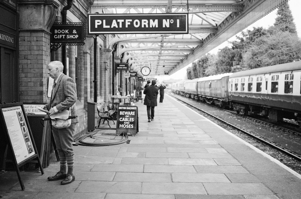 Looking down an historic train platform. On the tracks is a long set of carriages. On the platform is a man in an early 20th Century outfit. Further down the platform if a large sign declaring it to be "Platform No 1" and a man carries a small child. 
