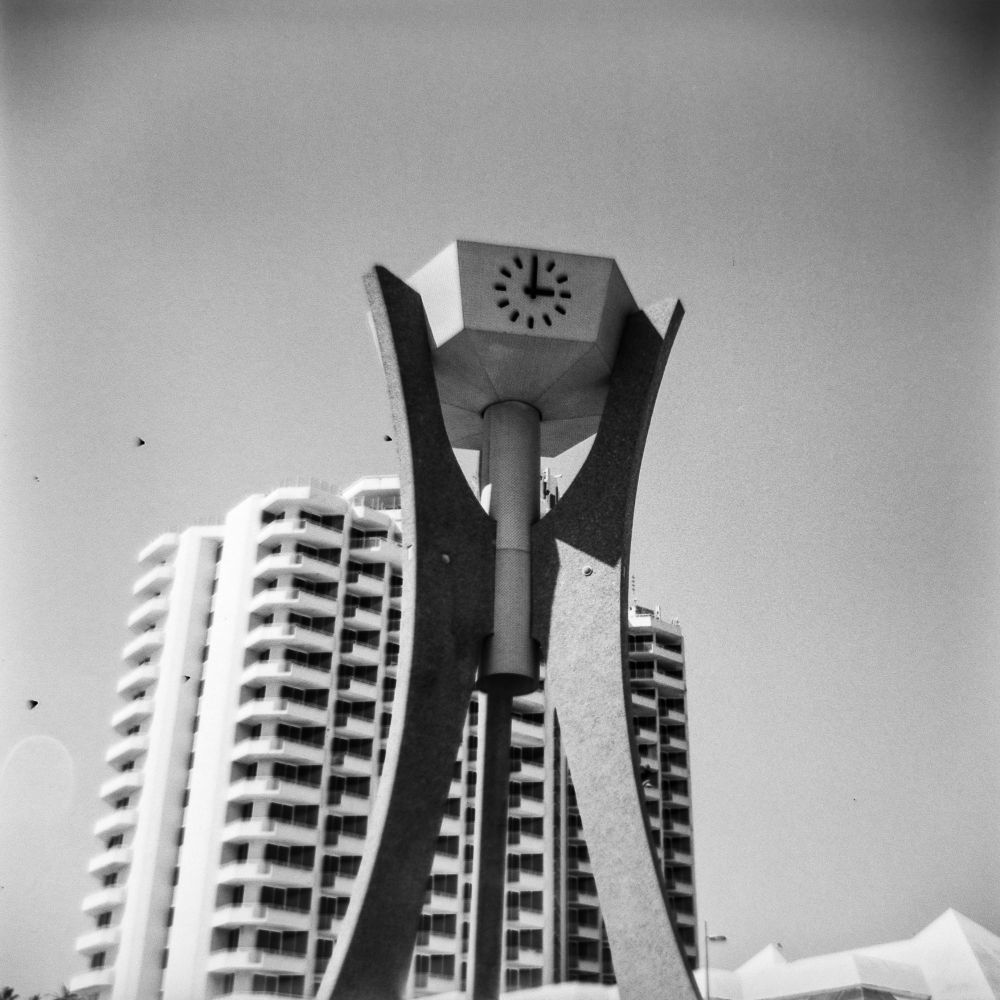 A black and white photo. A tall, concrete clocktower. The 3 legs are curved in a mid-century style, the clock on the top is a pentagonal shaped structure with a clock face. 
In the background is a very tall hotel building with about 15 floors. 