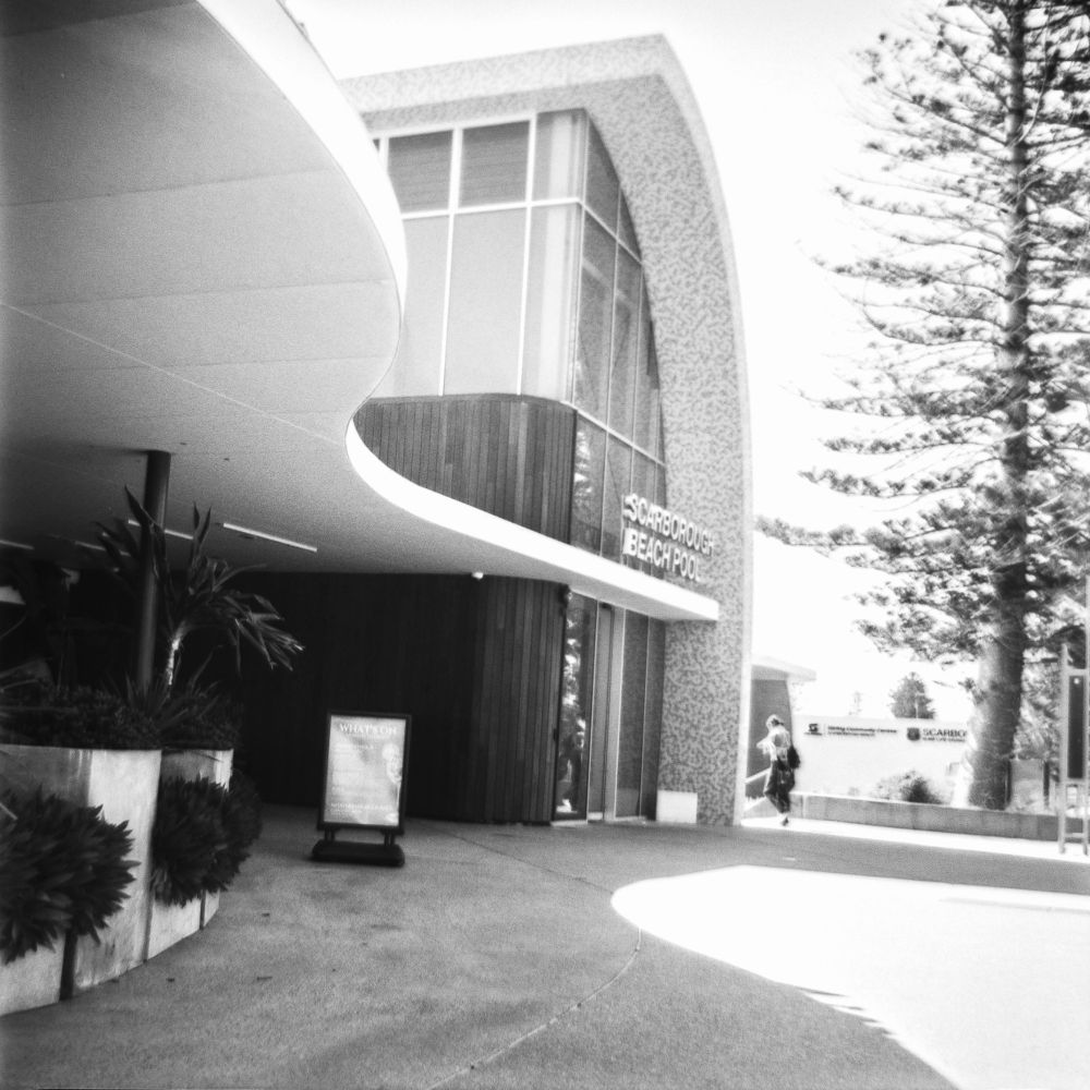 A black and white photo. The Scarborough Beach Pool building in a modern style. The roof in the left is flat and curved, the roof over the entrance is tiled and swoops upward. The entrance walls are all glass panels. 