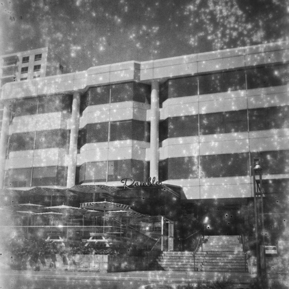 1990s era glass and concrete office block with an alfresco dining area in front. The dining area has striped umbrellas, and there are steps up to the entrance. The photo is black and white, and hazy because it is expired film. 