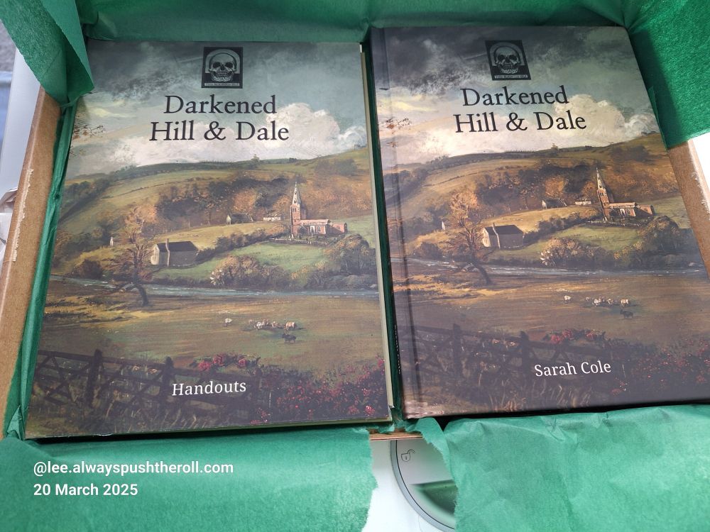 The just opened package of Darkend Hill & Dale, displaying the book and handouts pack, both nestled in green tissue paper.
They both depict the same artwork, a pastoral scene looking over a fence across a field with a small flock of sheep. In the middle distance is a river or large stream. Beyond the river are some buildings including a country church.