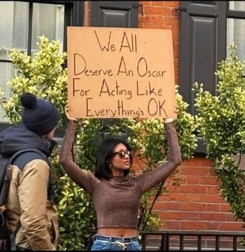 Woman in black sunglasses, stylish cropped long-sleeved brown tight sweater pulled up as she holds a sign the reads., We all deserve an Oscar for Acting like everything is Okay. Has lind of black panther, hipster intellectual funny badass feel.