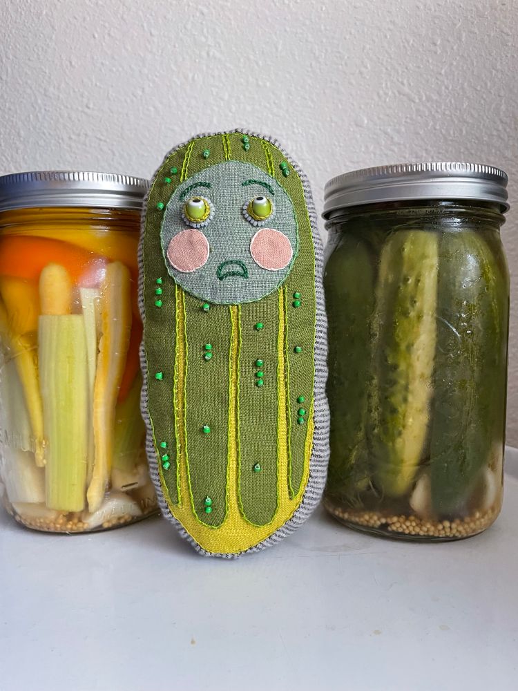 Hand sewn and embroidered pickle made of green linen with appliquéd stripes and beads to recreate pickle texture and a round face with round pink cheeks that is sighing and rolling its eyes in frustration. 