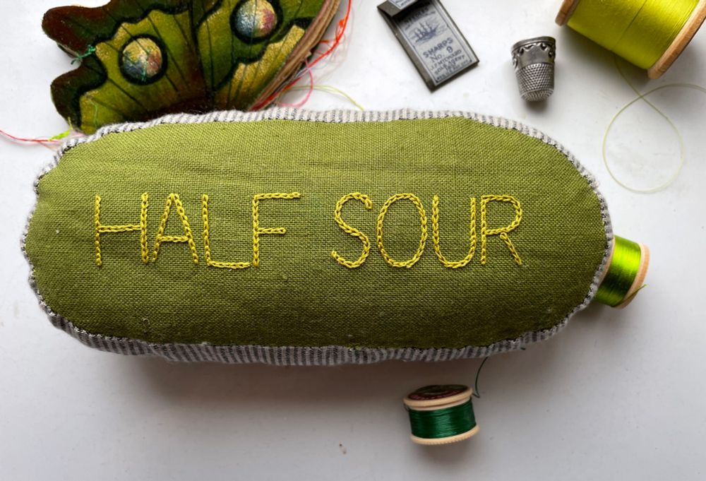 The back of a hand sewn, green linen pillow with the words HALF SOUR embroidered across it surrounded by sewing supplies. 
