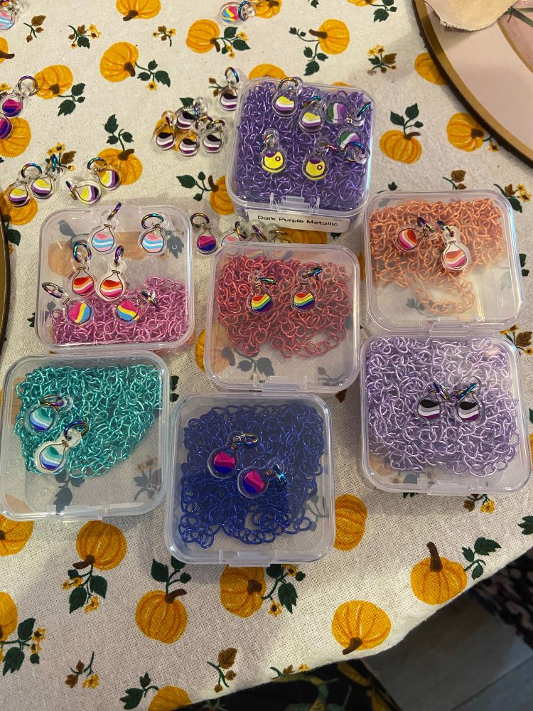 A collection of tiny charms shaped like potion bottles and the liquid is made up of colours of various LGBTQIA flag colours. There’s also boxes of colourful metal chains under them. 