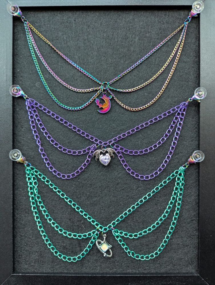 The triple layered mask chains pinned to a black pinboard. The top chain is made of a dark rainbow metal chain with a bat and moon charm in the centre. The middle one is made of a dark purple metallic chain with a purple heart with bat wings charm and the bottom is a sea green metallic chain with a 4 point star charm in the centre. 