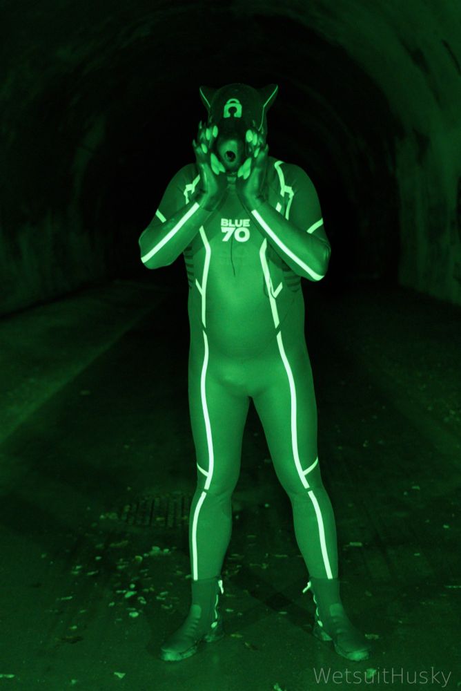 Person in a full-body BLUE70 wetsuit with white stripes and a Furrjoi drone mask stands in a dark tunnel, illuminated by green lighting. The scene evokes a rubber drone aesthetic.