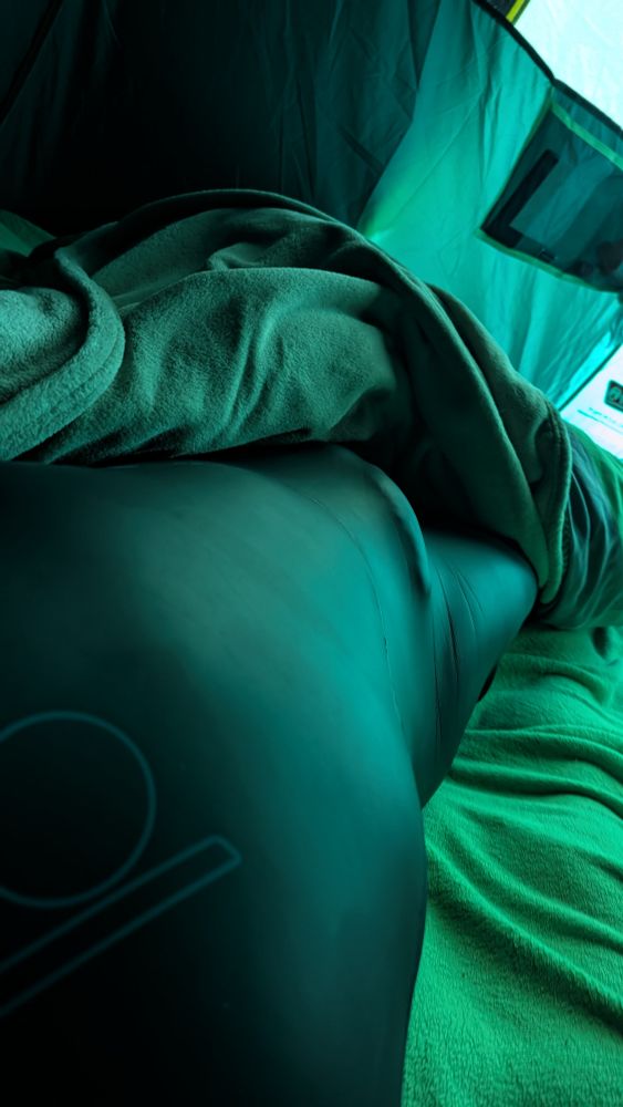 A person lying inside a tent, partially covered with a blanket, wearing a black wetsuit. The greenish light from the tent fabric gives the scene a teal tint.