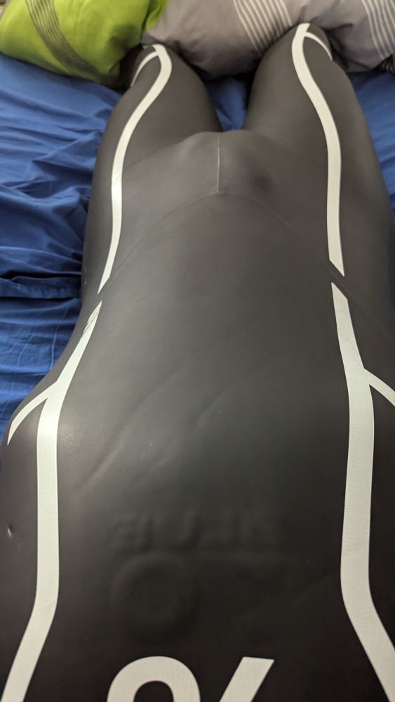 The image shows a person lying on a bed wearing a black neoprene wetsuit with white curved lines running down the legs. The wetsuit fits snugly, highlighting the contours of the body. The material has a smooth, slightly shiny texture, typical of neoprene, and there are a few visible creases in the fabric. The person is lying on their back with their legs together. The bed has blue sheets, and there are pillows in the background, including one that is green with a striped pattern. The perspective of the image is taken from above, looking down at the person’s body.