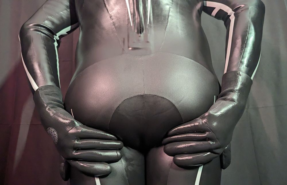 A close-up of the lower back and hips of a person wearing a tight black wetsuit with matte and glossy panels. They are wearing gloves and resting their hands on their hips. The background is dark.