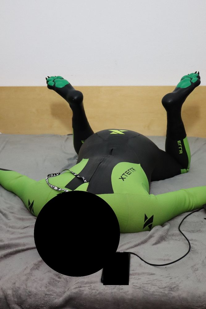 Person wearing a full-body black and green XTERRA wetsuit with paw-shaped feet, lying face down on a bed with legs bent upward in a relaxed, tired pose.