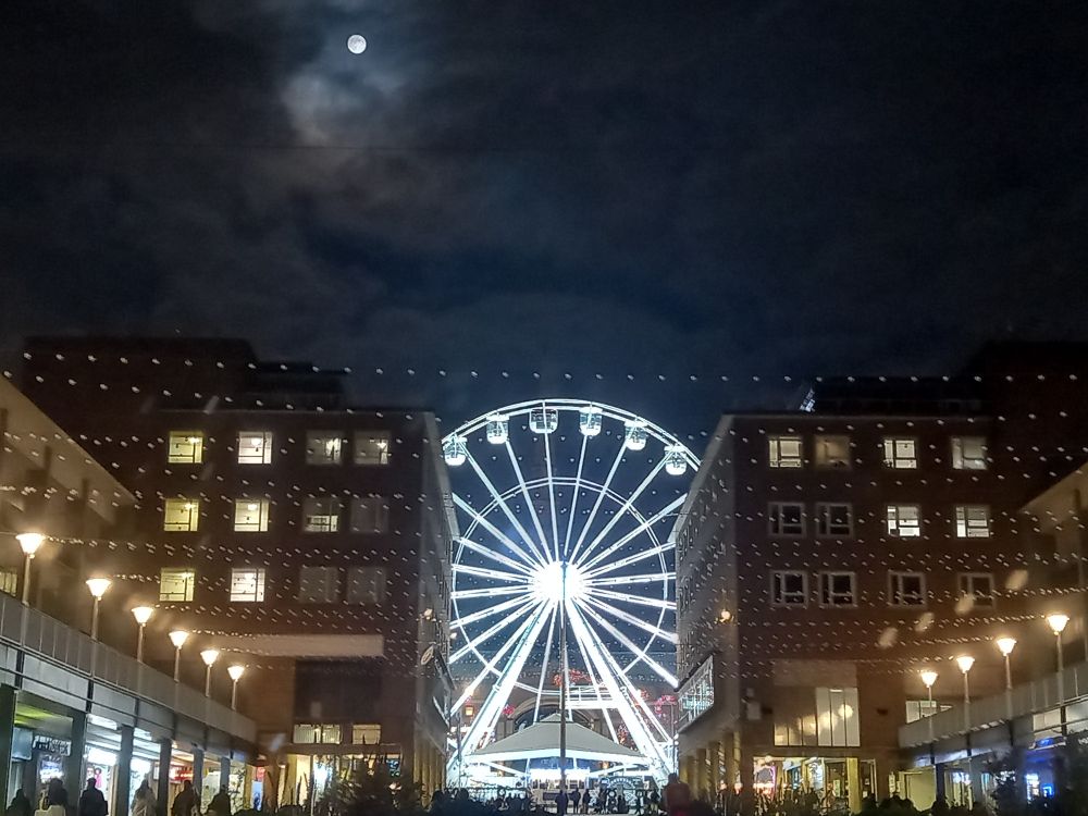 A city scene, taken in Coventry one night last year. A full moon in a cloudy sky hangs above a brightly lit ferris wheel, framed between buildings and shops.