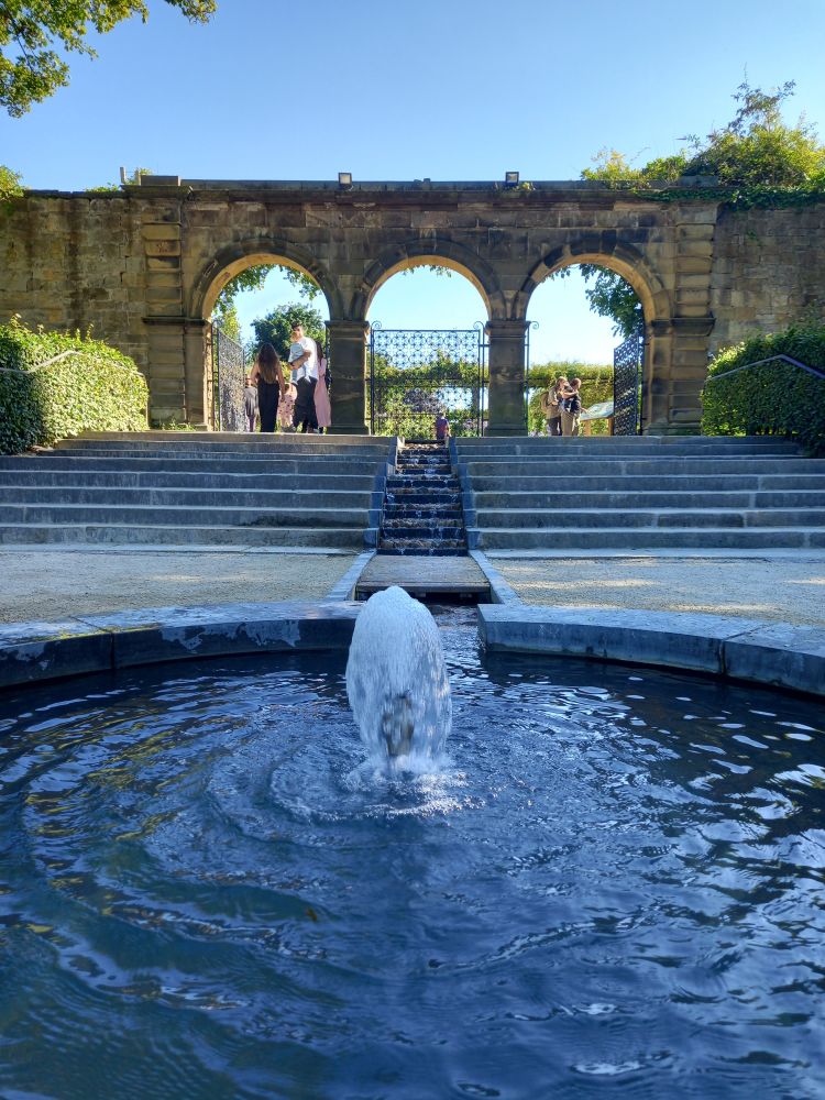 A fountain in a circular pool. Beyond, wide stone steps rise to the arched entrance into a formal garden. A clear blue sky lies beyond.