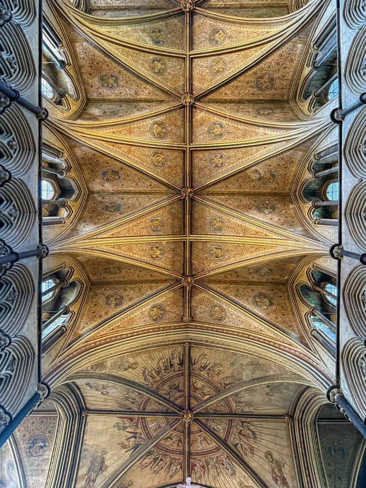 Ornate painted ceiling of Worcester Cathedral, viewed from directly below. The ceiling features golden ribbed vaulting with richly decorated panels containing floral patterns and medieval saintly figures. Toward the far end, a large circular mural depicts a celestial scene with angels radiating outward. Gothic arches and clerestory windows frame the view on both sides