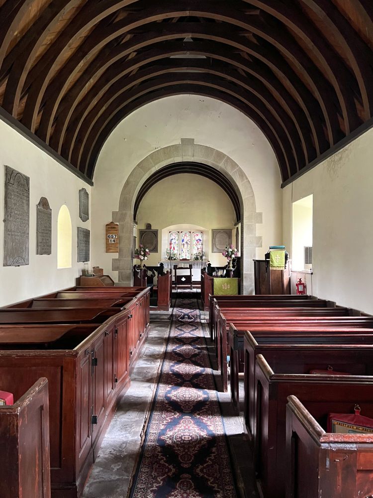 Interior of St Tegfedd’s Church in Llandegfedd — View down the central aisle toward the altar, framed by a stone arch. The church features a wooden barrel-vaulted ceiling, white plaster walls with memorial plaques, and rare early 19th-century wooden box pews on one side. A patterned carpet runner leads to the altar, which is adorned with floral arrangements and stained glass windows behind it. Soft natural light filters in through small arched windows, creating a serene atmosphere.