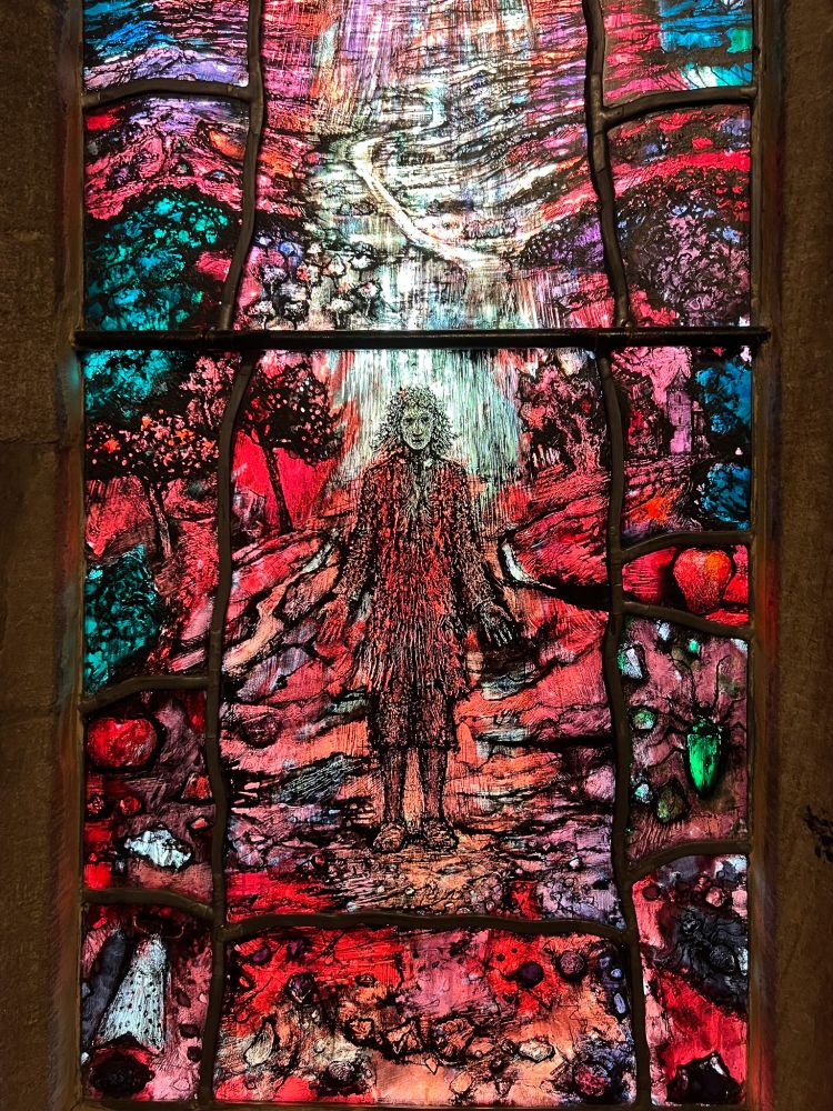 Stained glass window detail showing Thomas Traherne, in 17th century clerical dress, standing under a waterfall of light surrounded by aspects of creation.Colours are mainly red, with jewel-like effects of green, purple and turquoise.